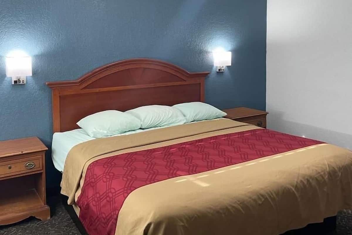A hotel room with a wooden headboard, a bed with a red and beige bedspread, two bedside tables, and wall-mounted lights.