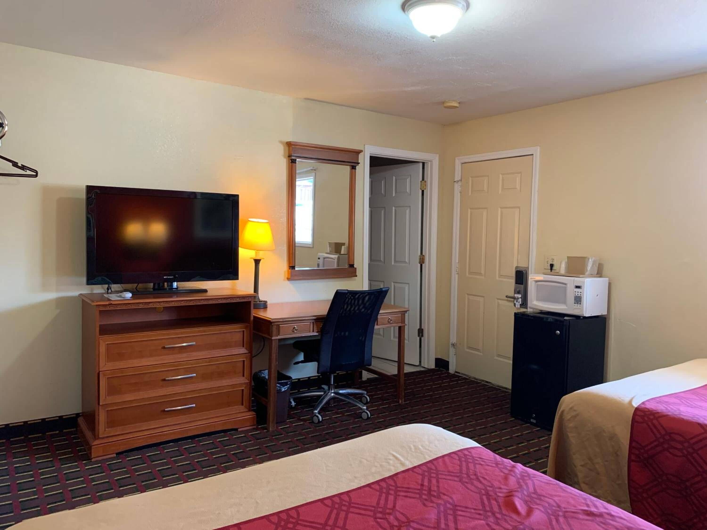 Una habitación de hotel con cama, escritorio, silla, televisor, lámpara, microondas y puerta.