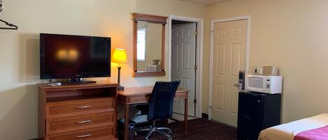 Una habitación de hotel con cama, escritorio, silla, televisor, lámpara, microondas y puerta.
