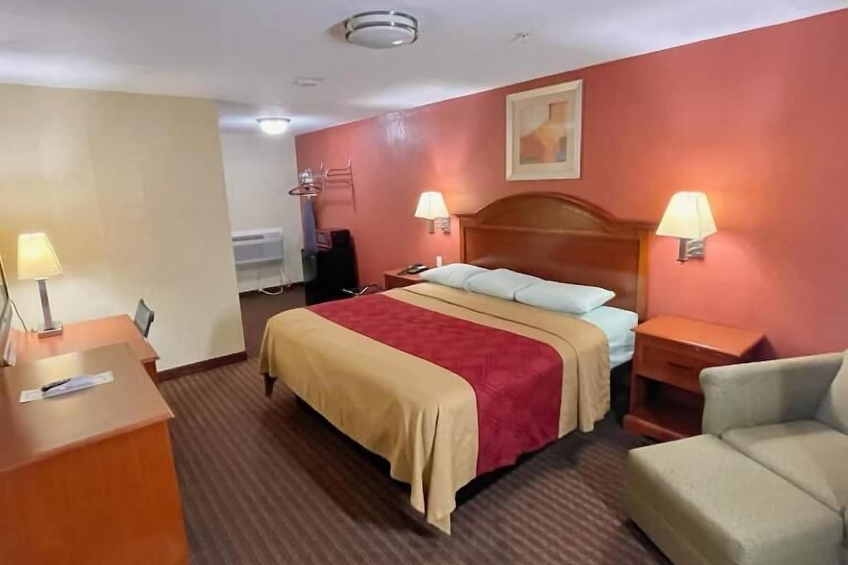 A hotel room with a bed, two bedside lamps, a desk with a chair, a waste bin, and a wall-mounted heater.