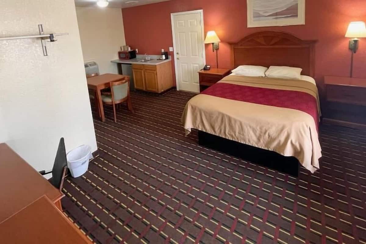 A hotel room with a bed, a desk, a chair, a small kitchenette, and a door.
