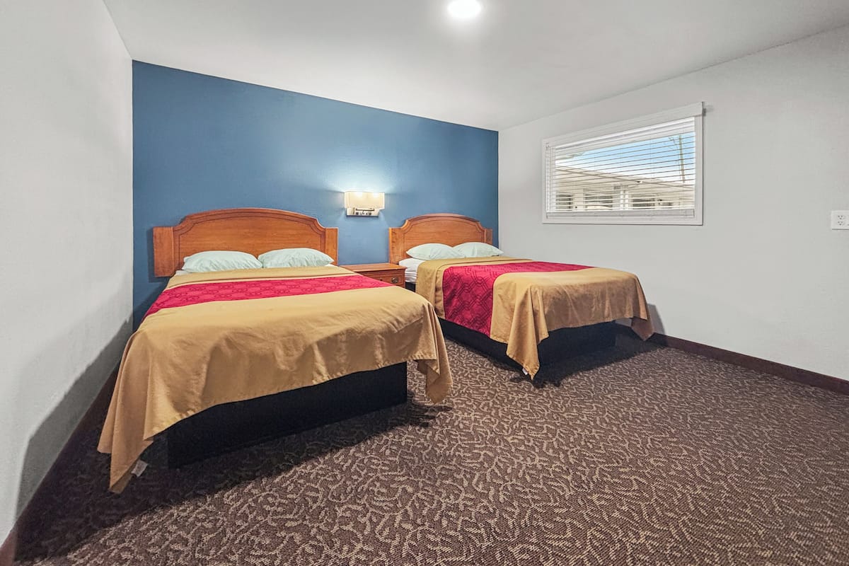 A hotel room with two beds, a blue accent wall, a window with blinds, and a carpeted floor.