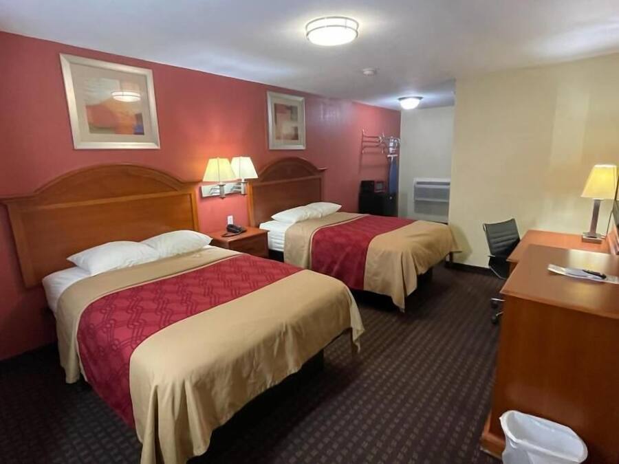 Ein Hotelzimmer mit zwei Betten, einem Schreibtisch, einem Stuhl, einem Mülleimer und einer Wandlampe.