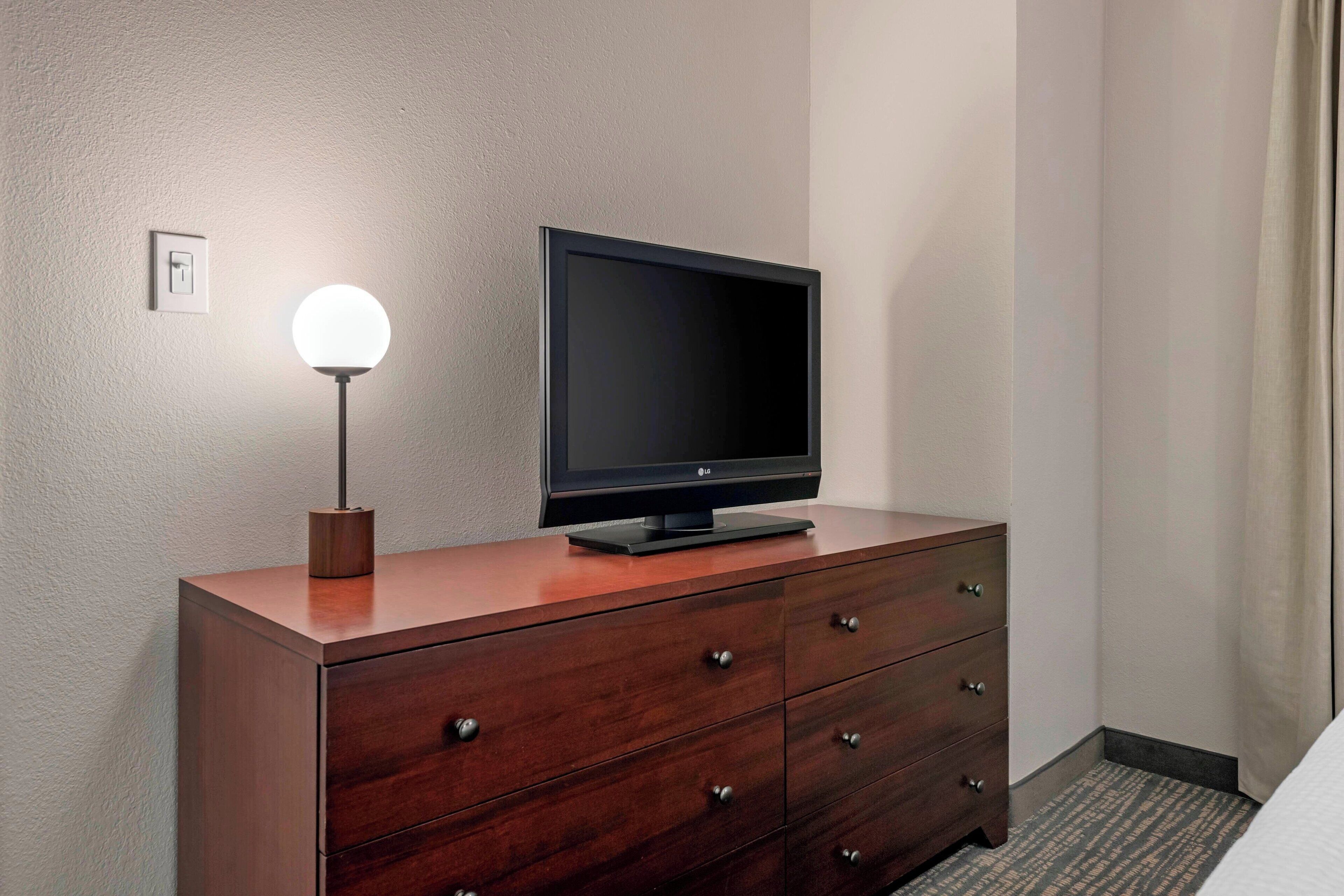 A hotel room with a wooden dresser, a flat-screen TV on top, and a wall-mounted light fixture.
