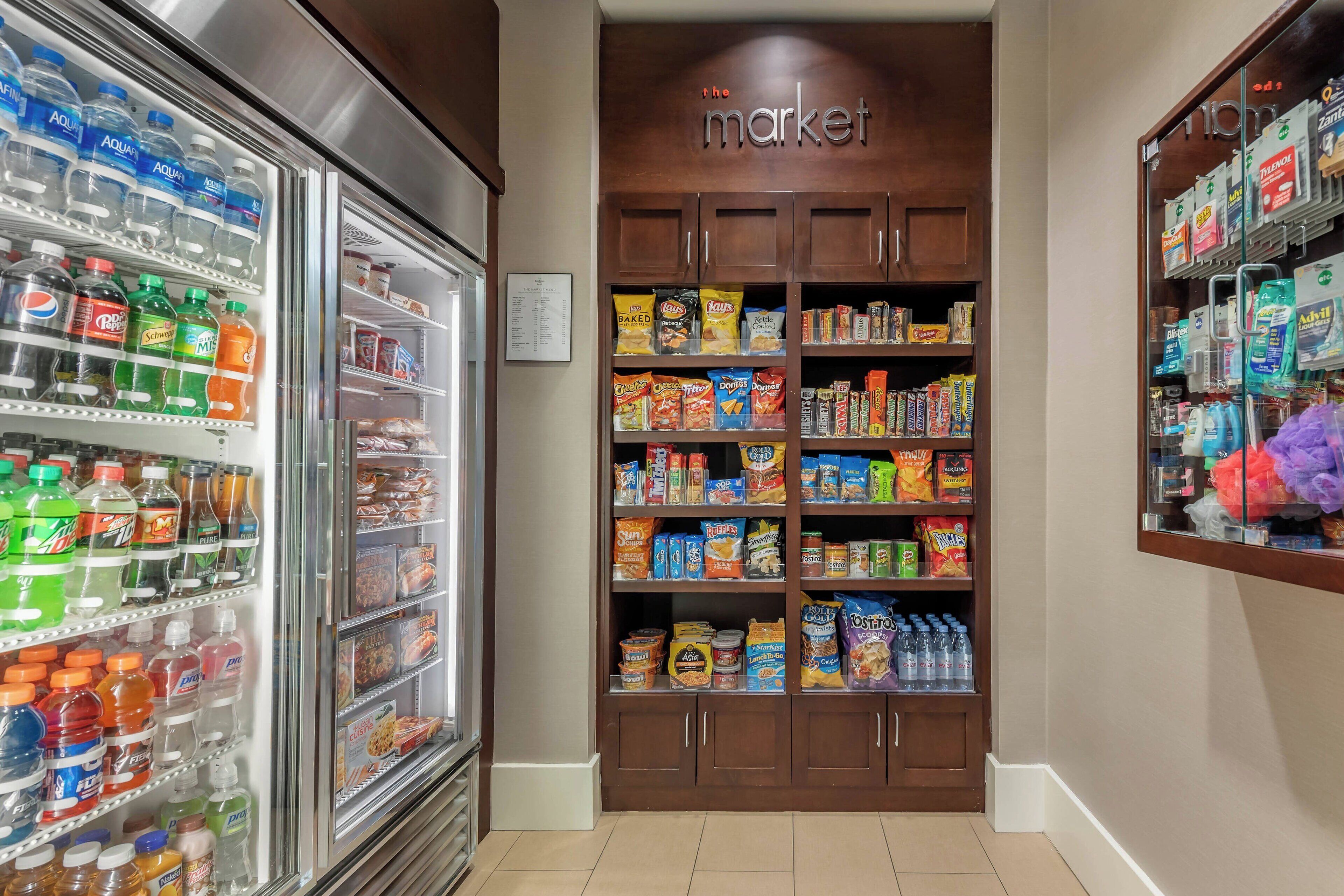 Interior de una tienda de comestibles con bebidas refrigeradas y estantes llenos de productos de snacks y artículos para el hogar.