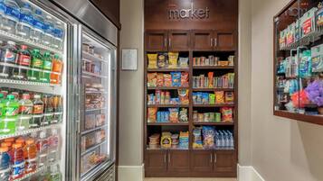 Interior de una tienda de comestibles con bebidas refrigeradas y estantes llenos de productos de snacks y artículos para el hogar.
