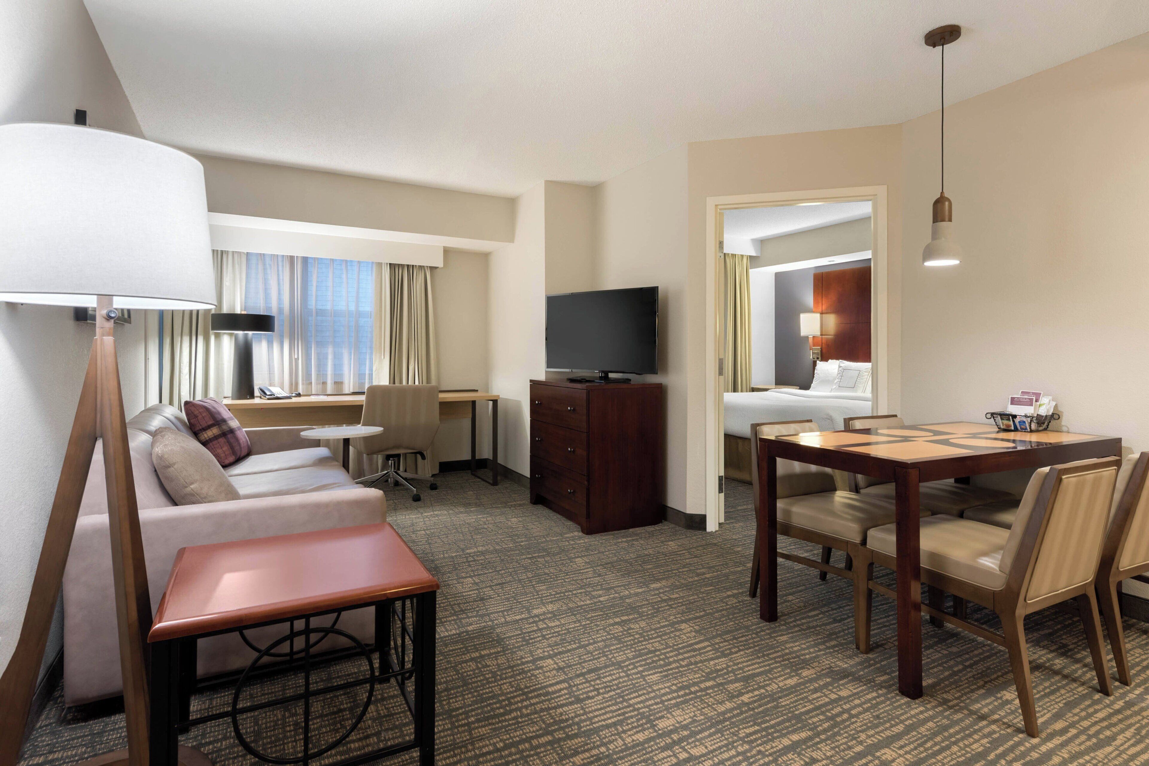 Una habitación de hotel con sofá, escritorio, televisión, mesa de comedor con sillas y lámpara.