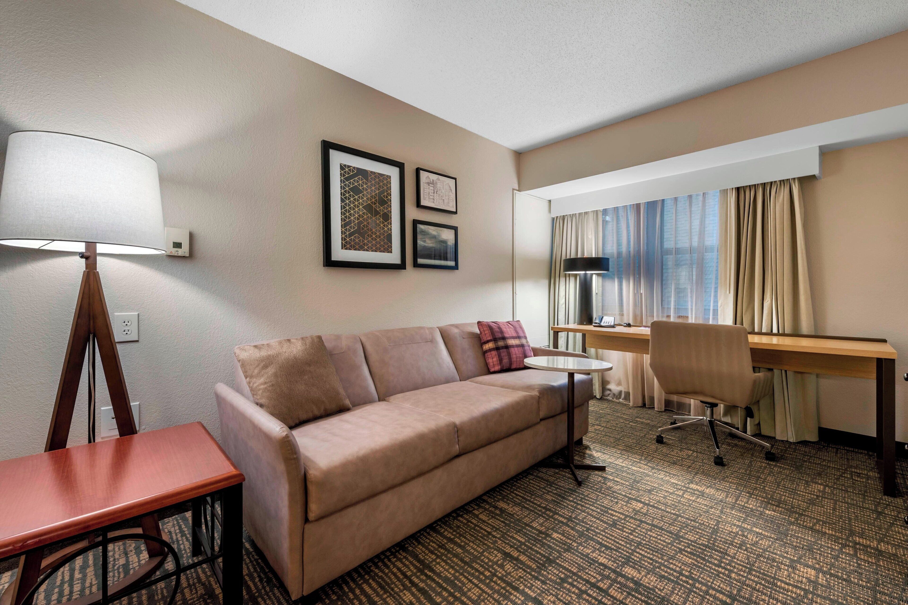 A hotel room with a sofa, a desk, a lamp, and a window with curtains.