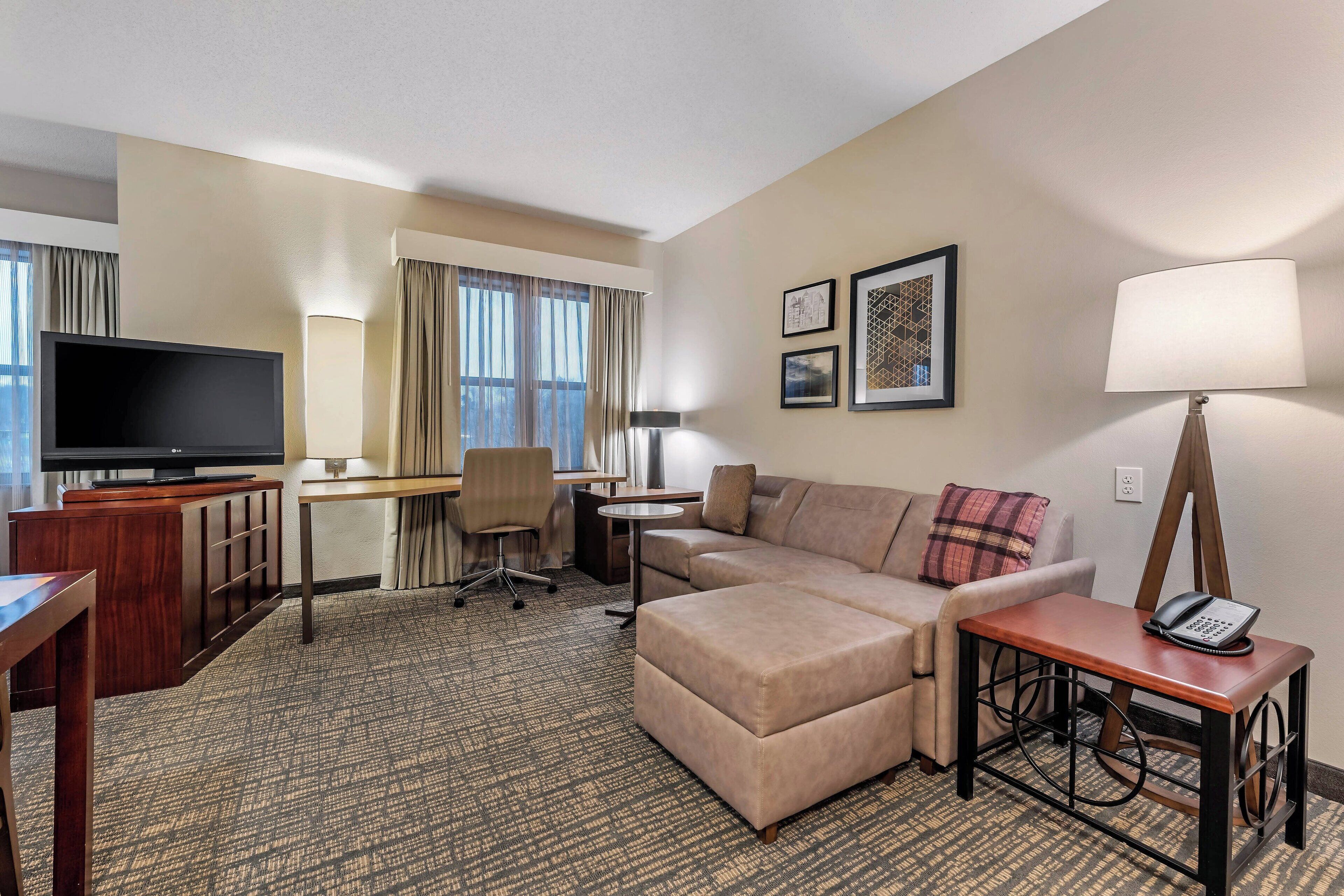 Quarto de hotel com sofá, escrivaninha, televisão, abajur e mesa pequena com telefone.