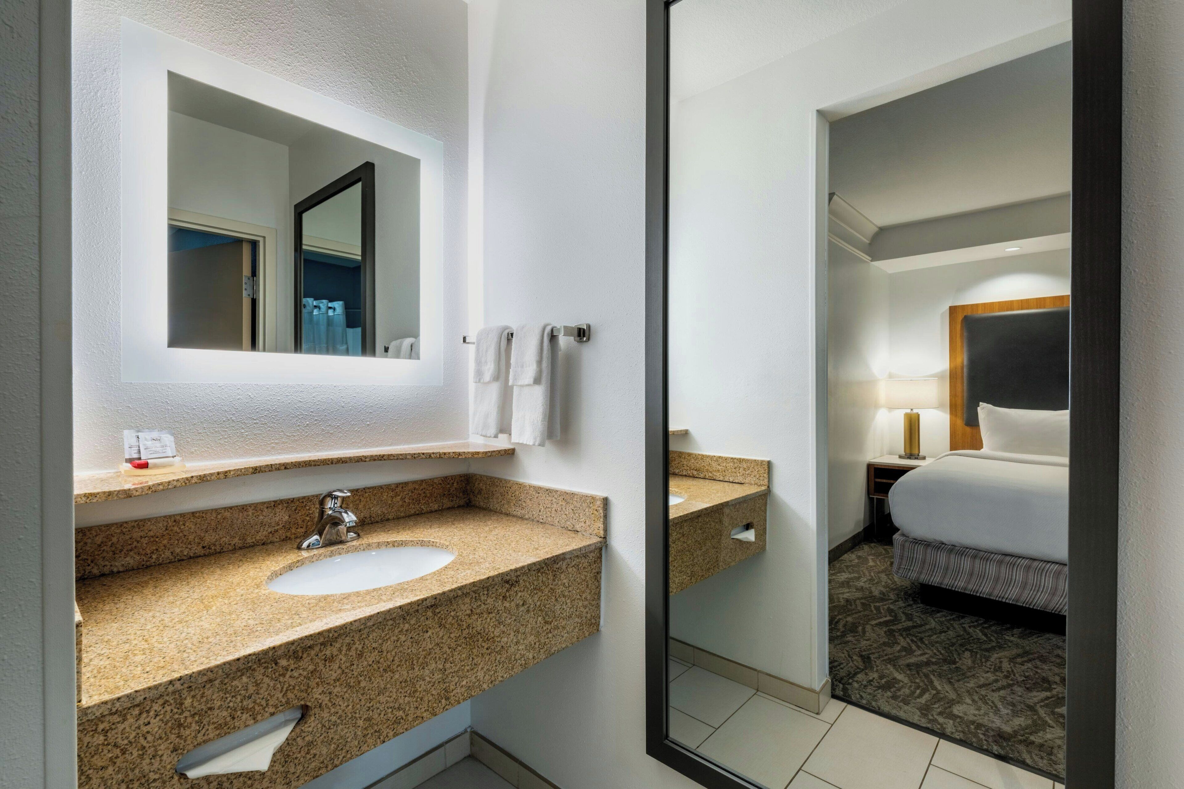 Ein Hotelbadezimmer mit Waschbecken auf einer Granitplatte, einem großen Spiegel und Blick auf ein Schlafzimmer mit Bett und Nachttisch.