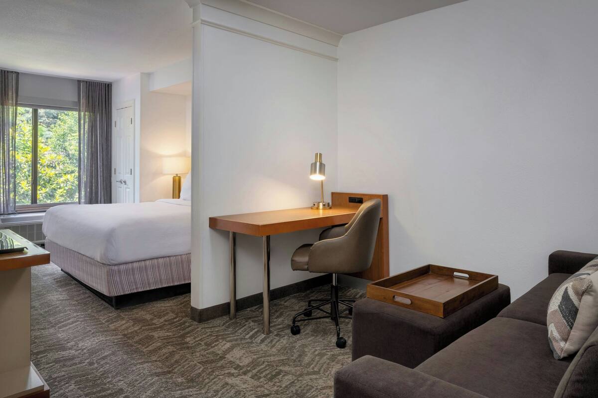 Una habitación de hotel con cama, escritorio, silla, sofá y ventana con cortinas.