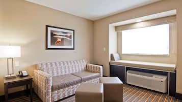 A hotel room with a sofa, ottoman, bedside table with a lamp, a framed picture on the wall, a window with a view, and an air conditioning unit.