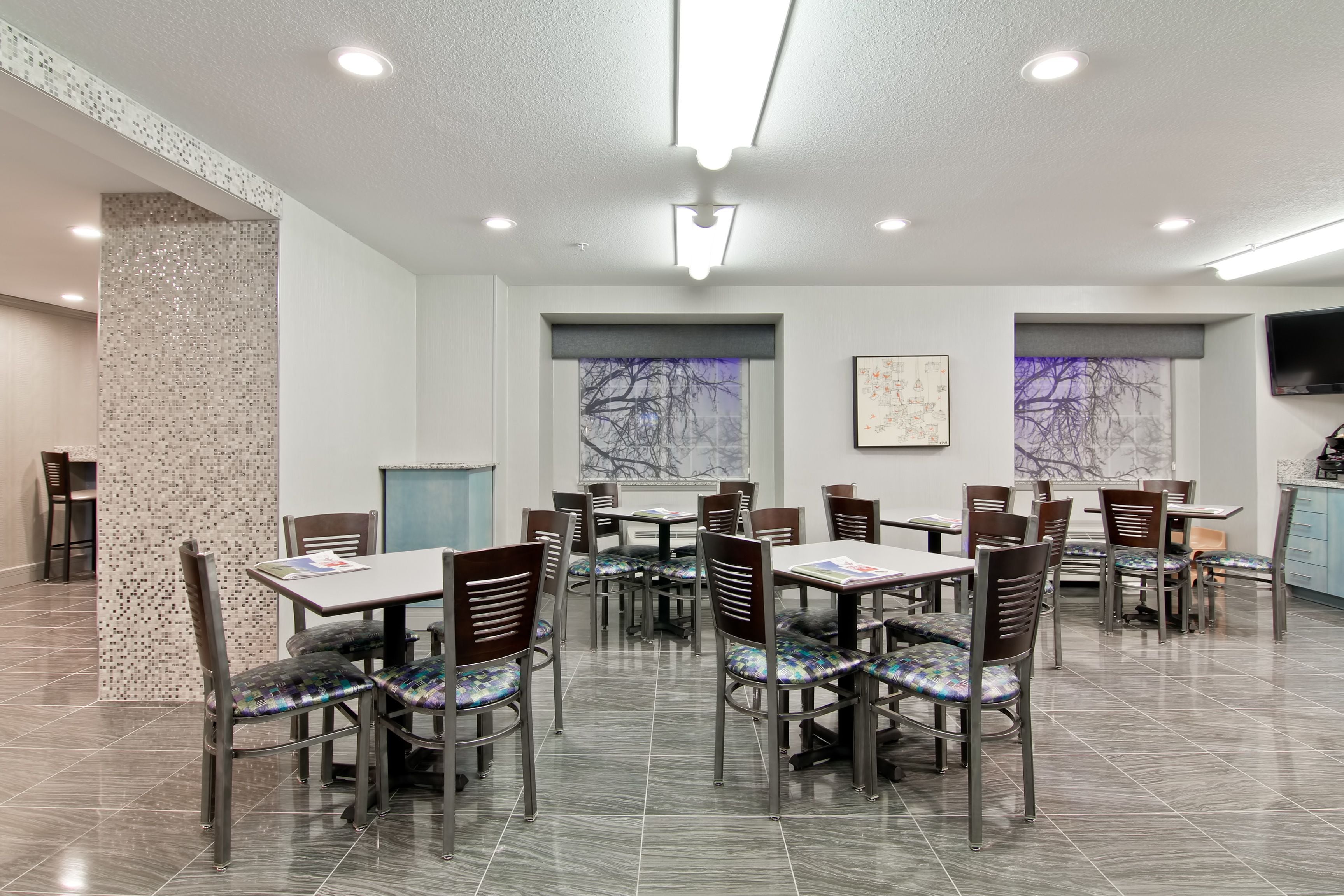Uma sala de jantar com várias mesas e cadeiras, chão de azulejos e uma grande janela com vista para árvores sem folhas.