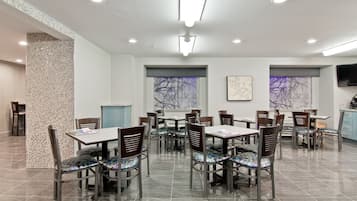 Uma sala de jantar com várias mesas e cadeiras, chão de azulejos e uma grande janela com vista para árvores sem folhas.