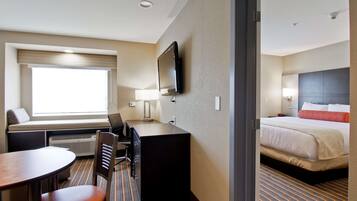 A hotel room with a bed, a desk with a chair, a television mounted on the wall, and a window with blinds.