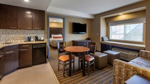 A compact hotel room with a kitchenette, a dining area with a round table and chairs, a bed with a TV above it, and a window with a view.