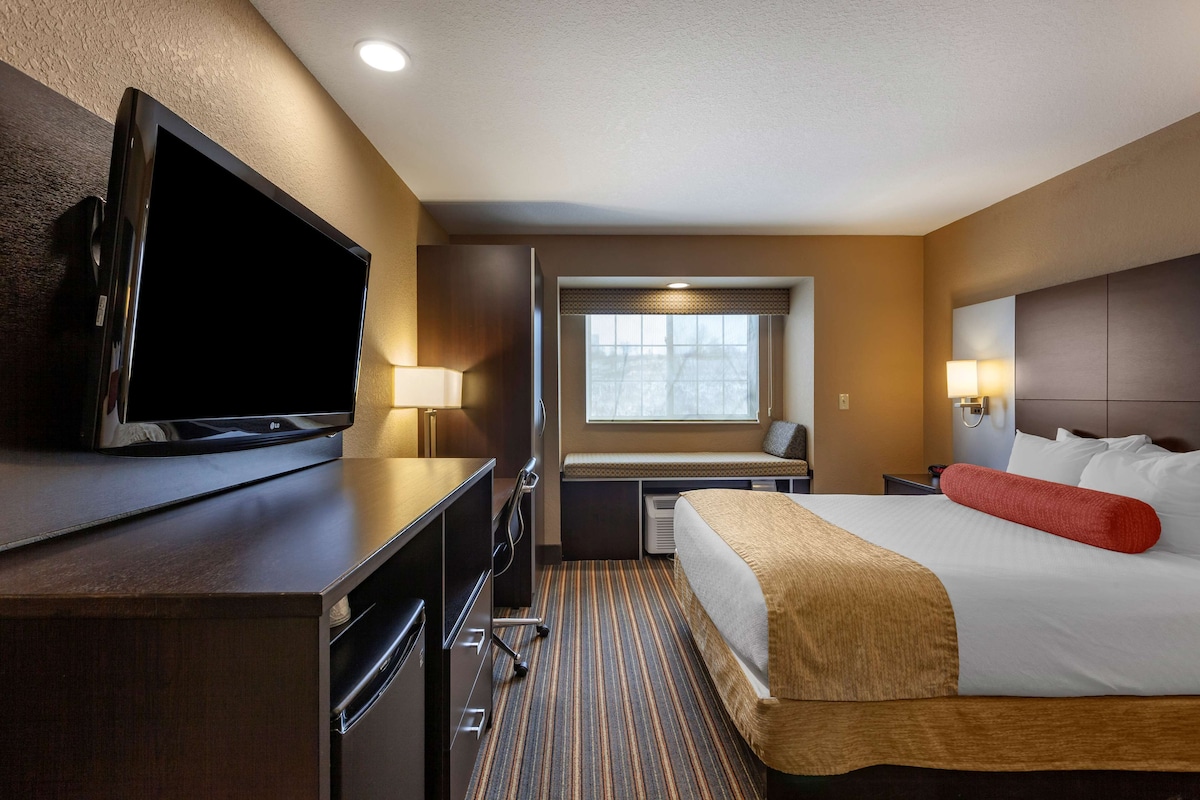 A hotel room with a bed, a desk with a flat-screen TV, a window with blinds, and a lamp.