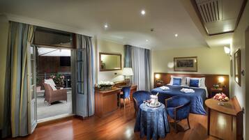 A hotel room with a bed, a desk, a chair, a small table, and a balcony with a wicker chair and flowers.