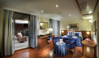 A hotel room with a bed, a desk, a chair, a small table, and a balcony with a wicker chair and flowers.
