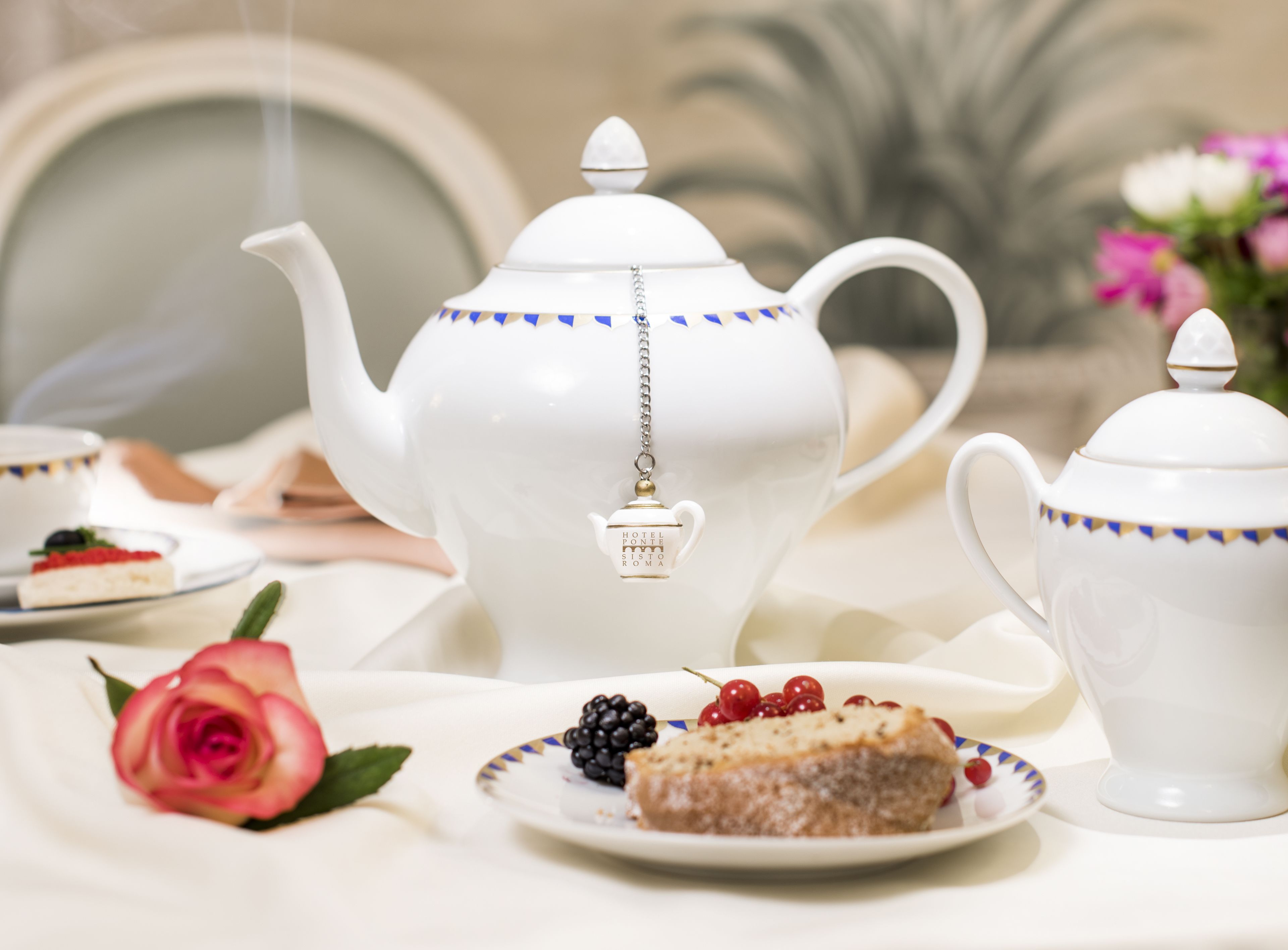 A tea set with a teapot and cups, a slice of cake with berries, and a rose on a table.