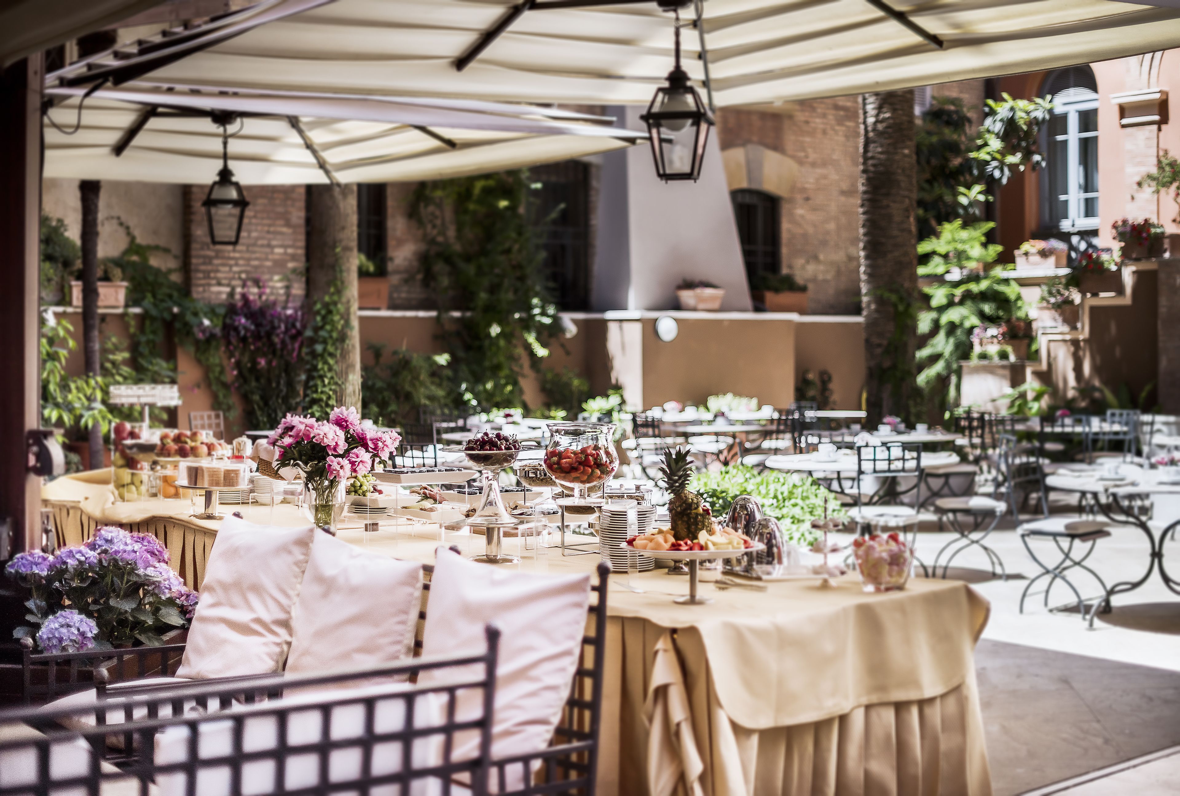 An outdoor dining area with tables set for a meal, surrounded by plants and a brick wall.