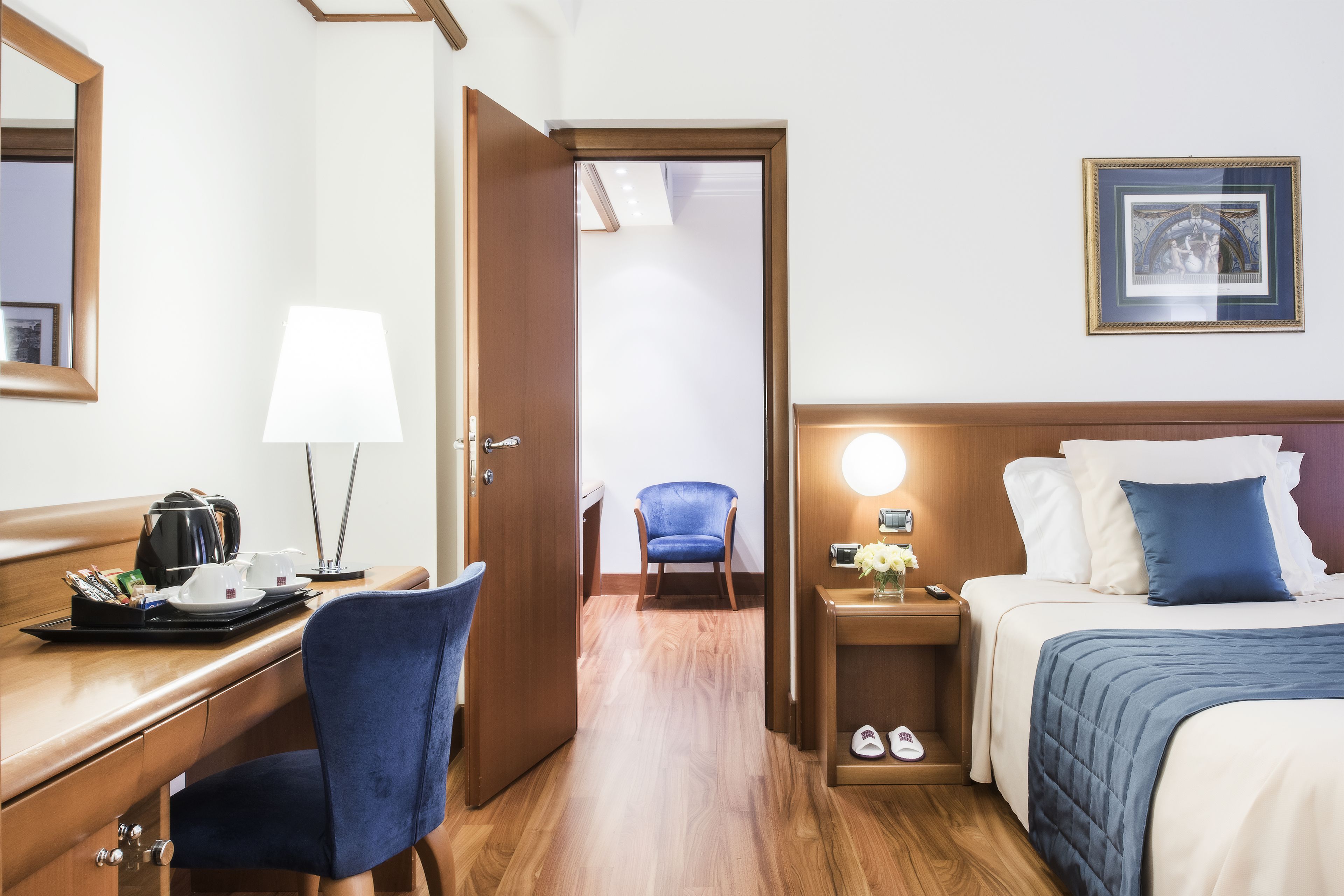 Une chambre d’hôtel avec un bureau en bois, une chaise, un lit avec un oreiller bleu, une table de chevet avec une lampe et un tableau encadré au mur.