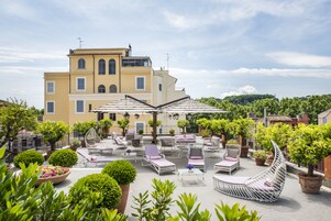 A rooftop terrace with outdoor seating, potted plants, and a building in the background.