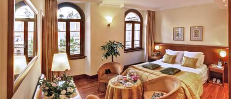 A hotel room with a bed, two armchairs, a round table with a vase of flowers, a lamp, and a nightstand with a clock.