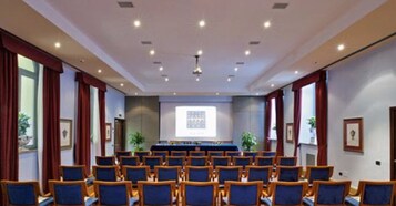 Una sala conferenze con file di sedie, uno schermo di proiezione e un’area per presentazioni.