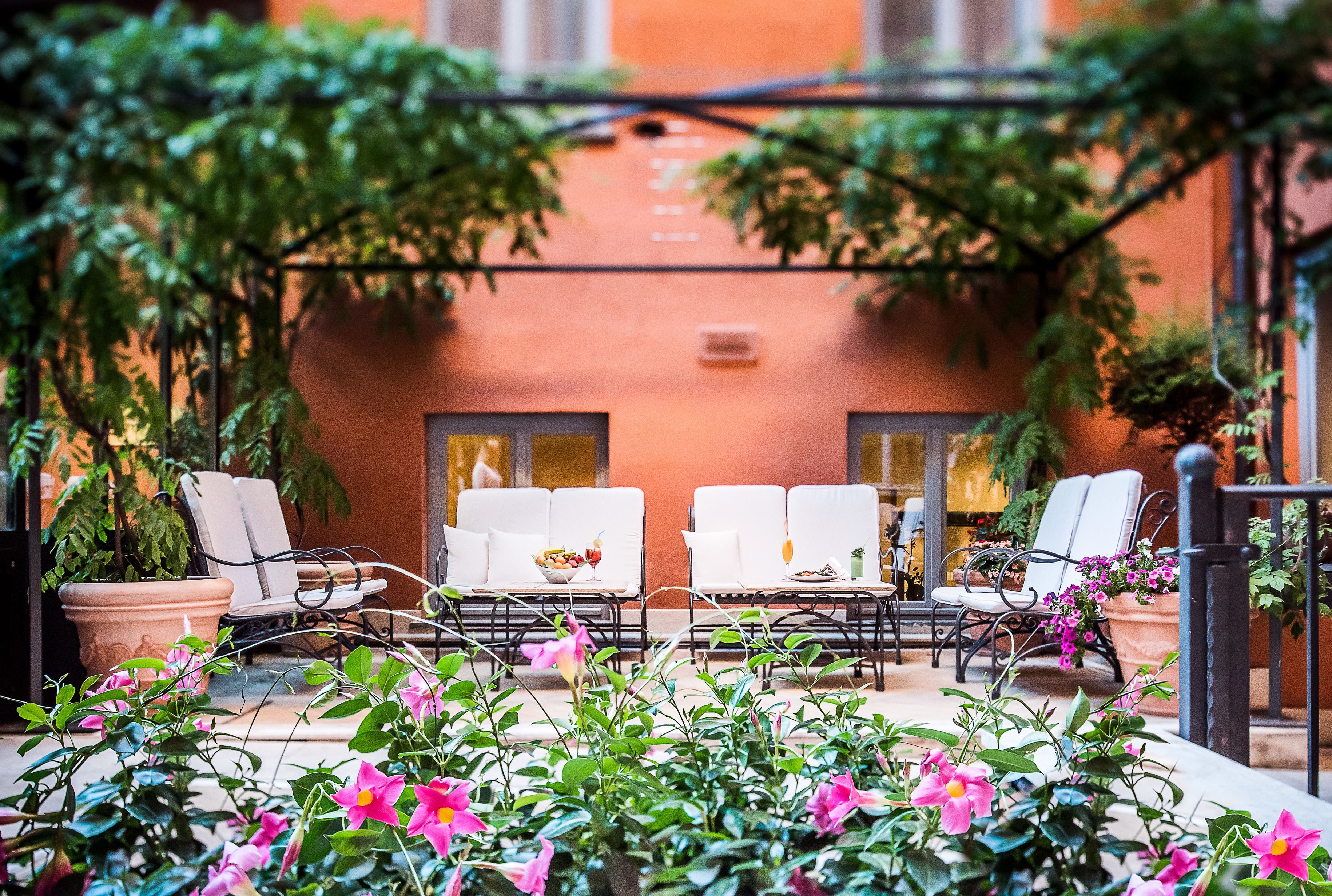 An outdoor seating area with white chairs and terracotta pots, surrounded by lush greenery and pink flowers.