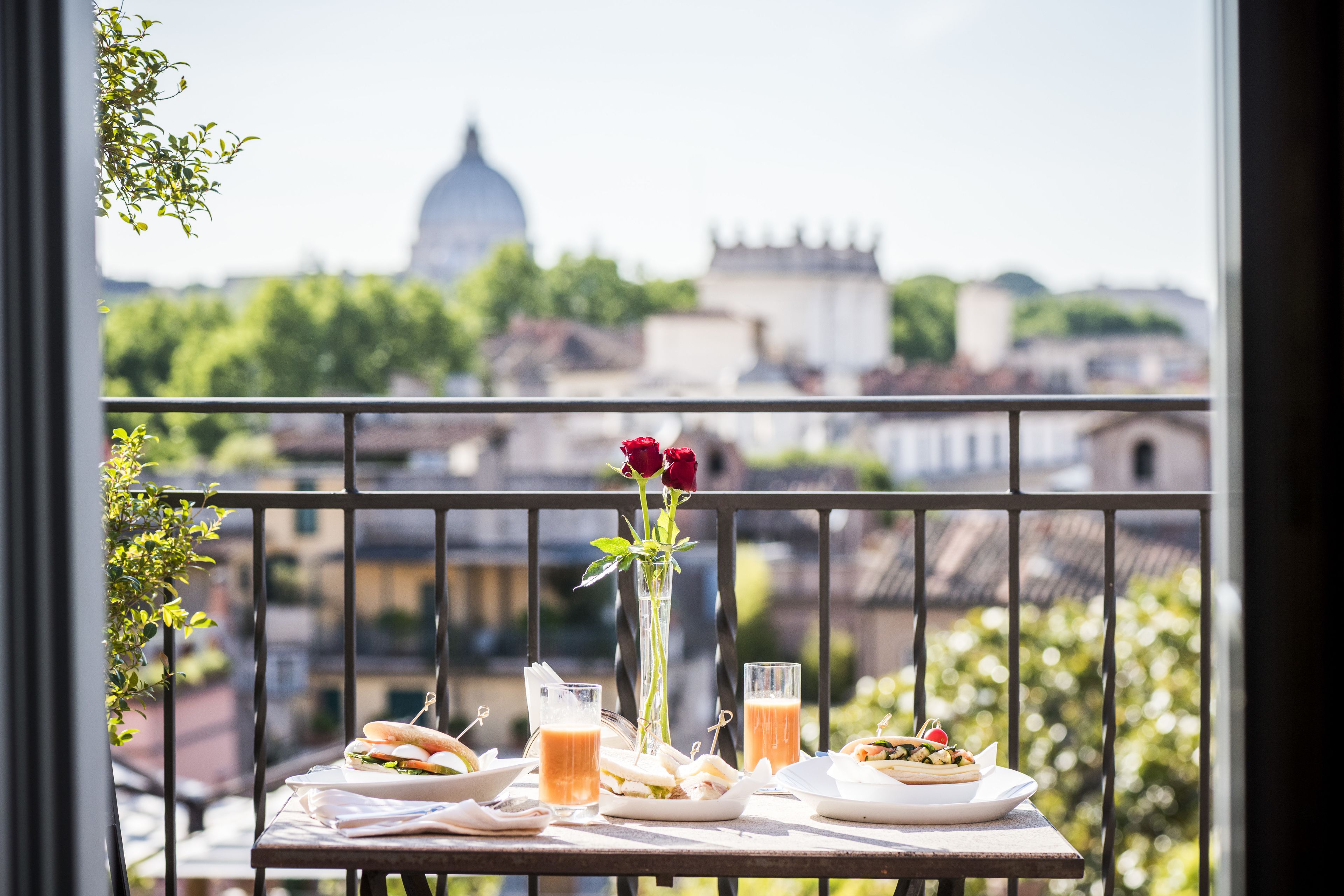 A balcony with a table set for lunch, a vase with a red rose, and a view of a cityscape including a dome and buildings.