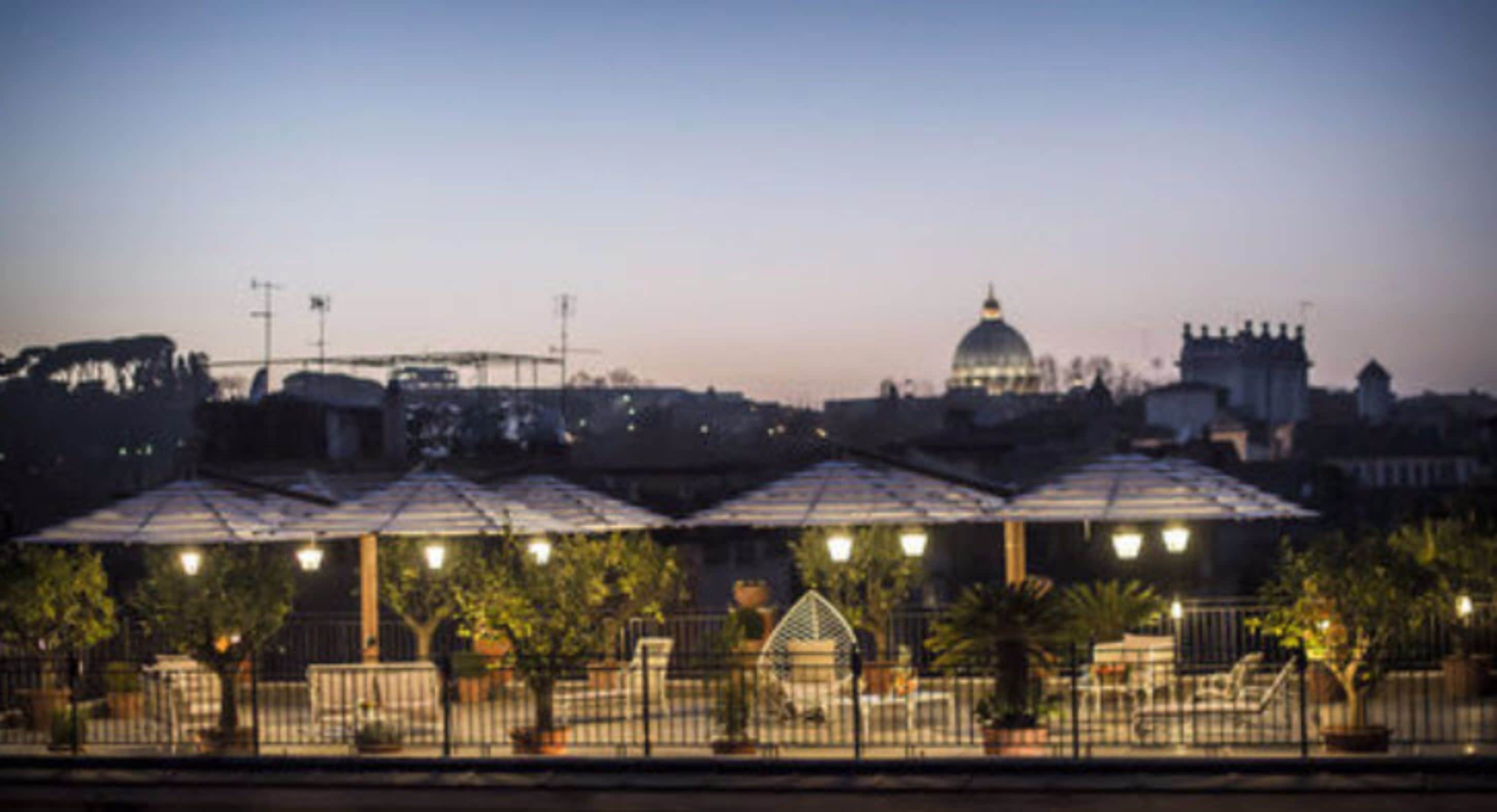 A nighttime view of a cityscape with a prominent dome structure in the background, a series of covered outdoor seating areas in the foreground, and a body of water separating the seating area from the city.