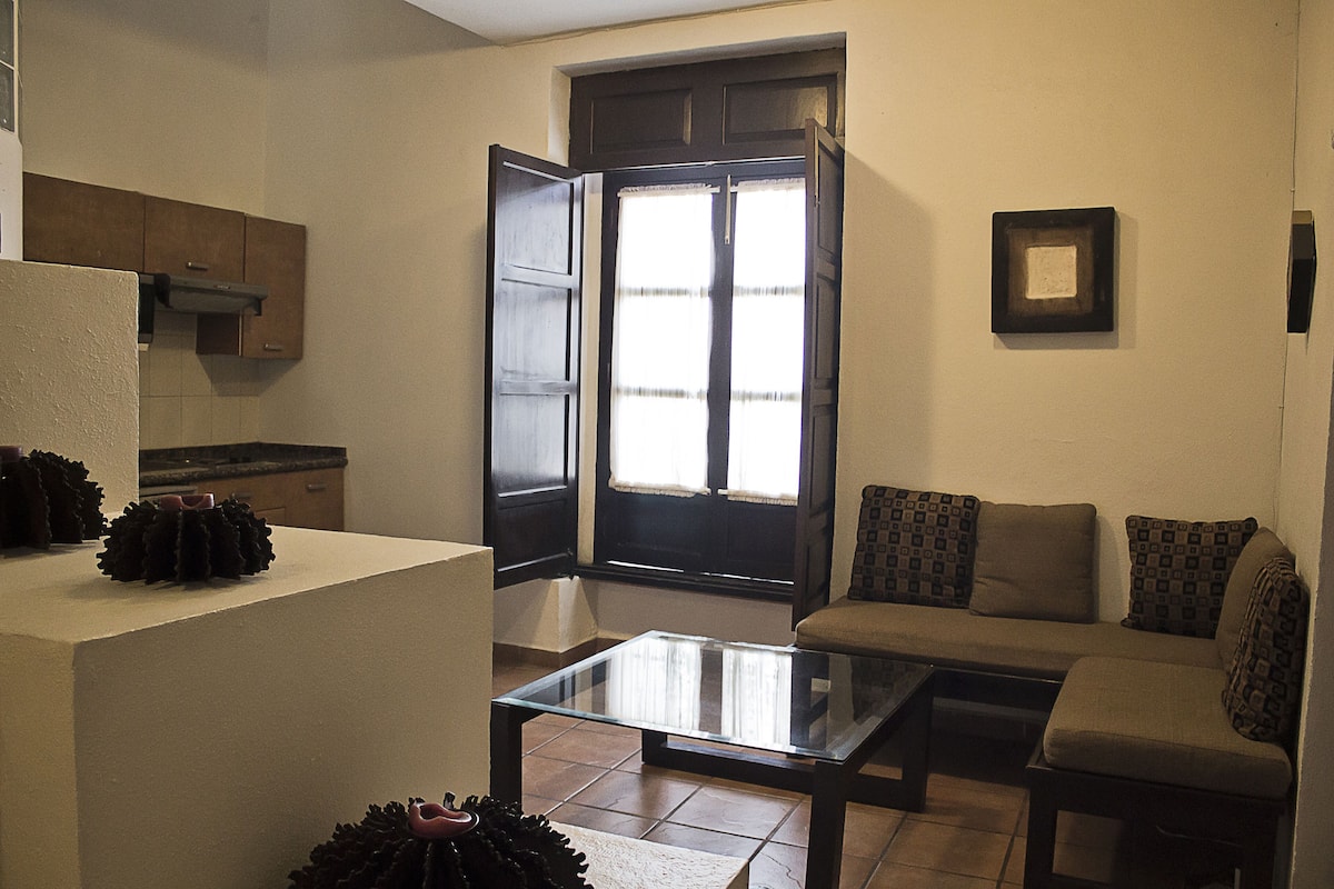 A compact living space with a glass coffee table, a sofa, a kitchen area, and a large window.