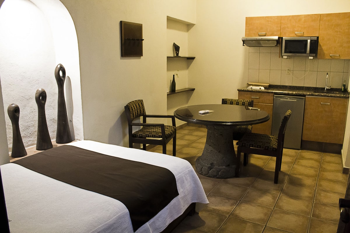 A compact hotel room with a bed, a dining table, a chair, a small kitchen area with a microwave and a dishwasher, and a tiled floor.