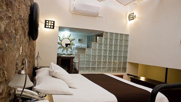 A hotel room with a bed, bedside lamp, phone, and a small table with a lamp. There is a brick wall on one side and a tiled wall on the other. - Hotel Suites Sofia (Querétaro)