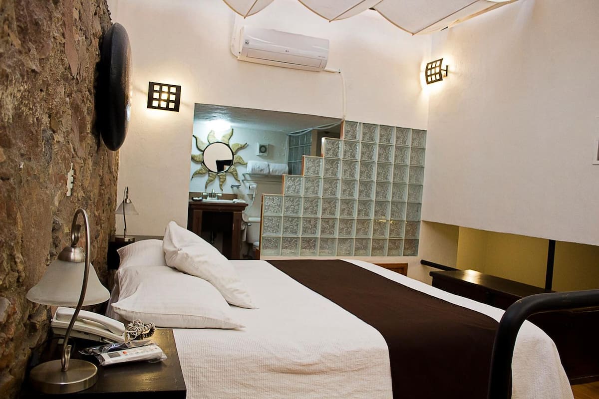 A hotel room with a bed, bedside lamp, phone, and a small table with a lamp. There is a brick wall on one side and a tiled wall on the other.