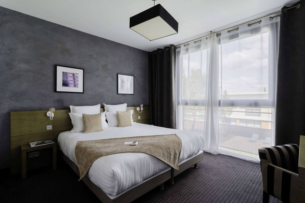 Een moderne hotelkamer met een groot bed, een nachtkastje, een stoel en uitzicht op de omgeving door dunne gordijnen.