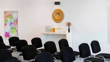 Une salle de conférence avec des chaises noires disposées en rangées, un comptoir d’accueil blanc agrémenté d’une décoration murale, et un tableau abstrait aux couleurs vives accroché au mur.