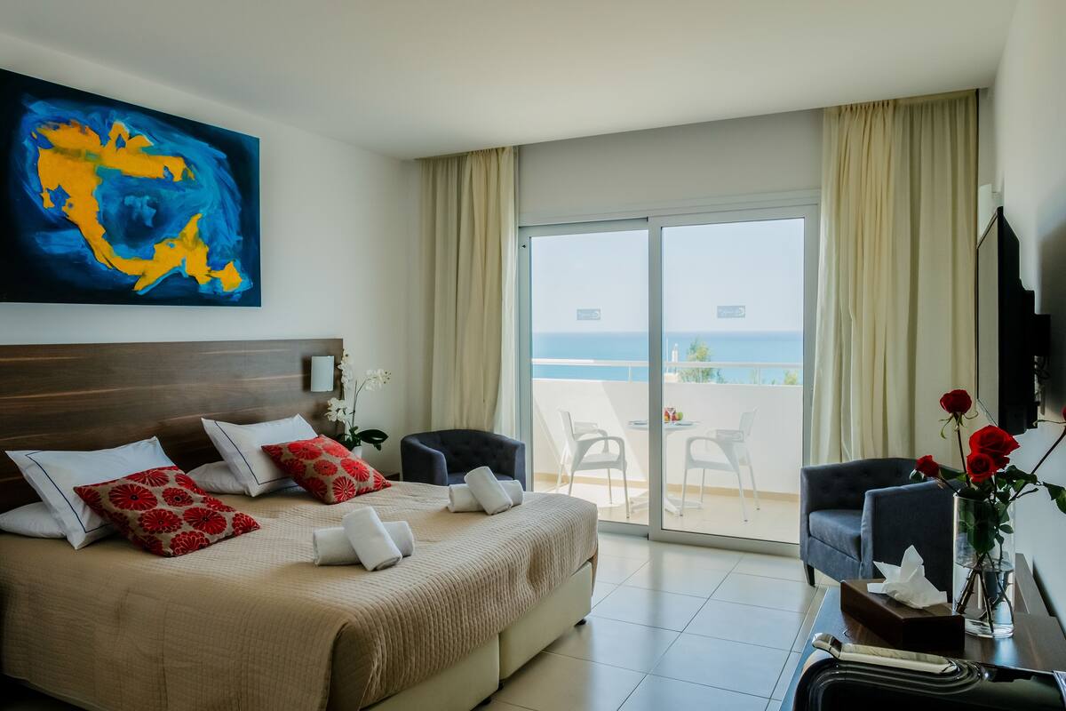 Une chambre d’hôtel avec un lit, un bureau agrémenté d’un vase de fleurs, une chaise et une grande fenêtre donnant sur la mer.