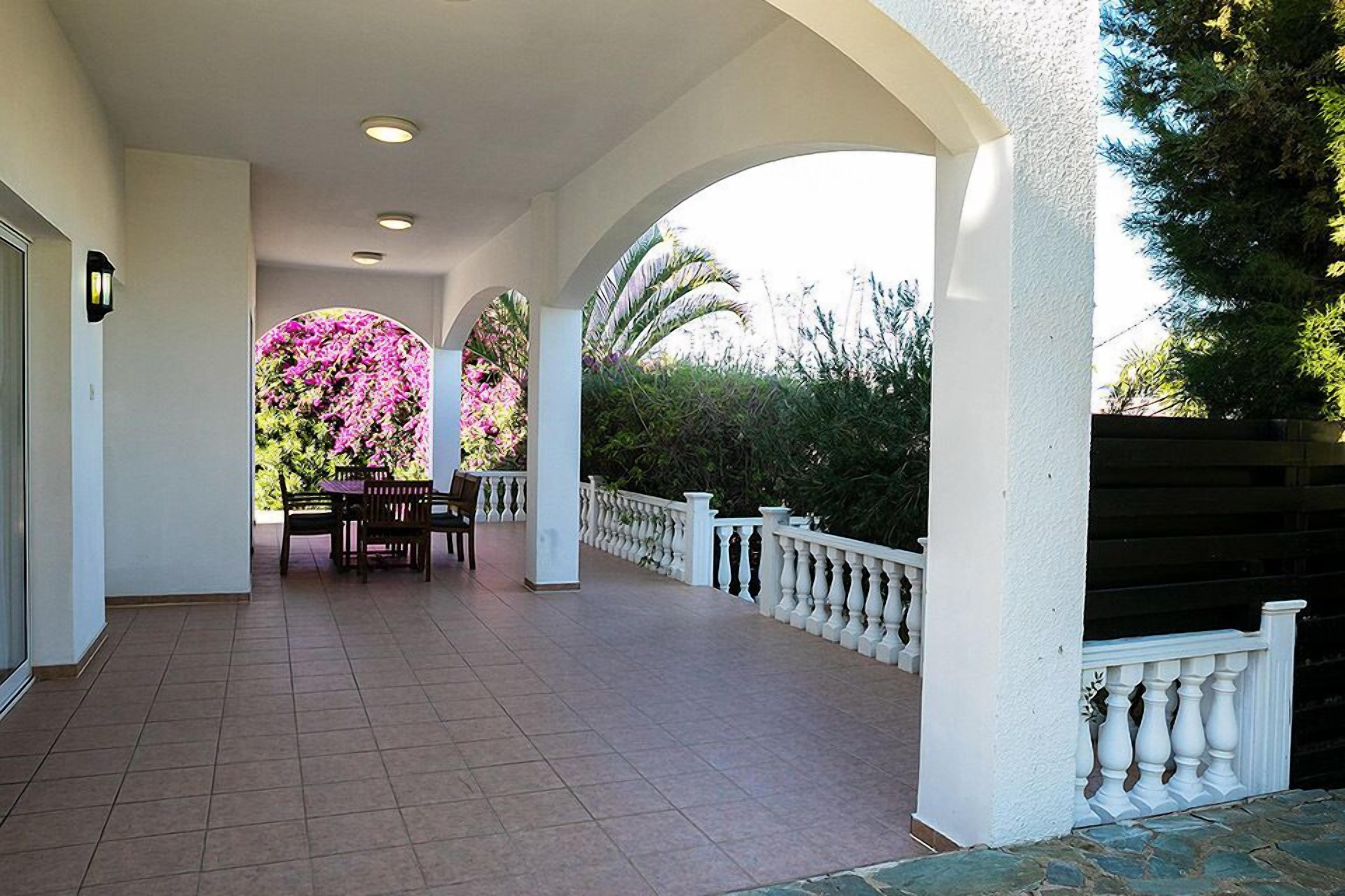 A covered outdoor area with a table and chairs, surrounded by white railings and greenery.