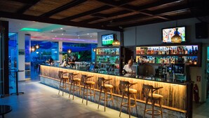 A bar area with wooden bar stools, a counter with a TV above, and shelves stocked with bottles. - Theo Sunset Bay Hotel (Kissonerga)