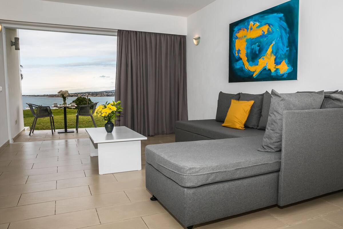 Un salon moderne avec un canapé gris, une table basse blanche et une vue sur la mer grâce à de grandes fenêtres.