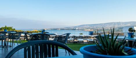 An outdoor seating area with tables and chairs, overlooking a body of water and distant hills.