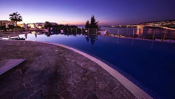 A nighttime pool area with a clear view of the water reflecting the surroundings, a stone deck, and distant city lights. - Theo Sunset Bay Hotel (Kissonerga)