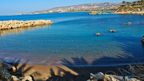 A coastal scene with clear blue waters, rocky shore, and a distant hilly landscape. - Theo Sunset Bay Hotel (Kissonerga)