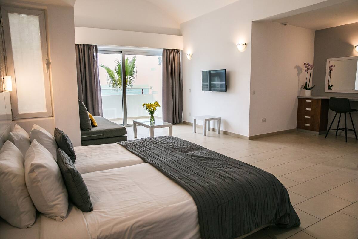 Une chambre d’hôtel avec un lit, un bureau, une chaise, une télévision et un balcon avec des rideaux.