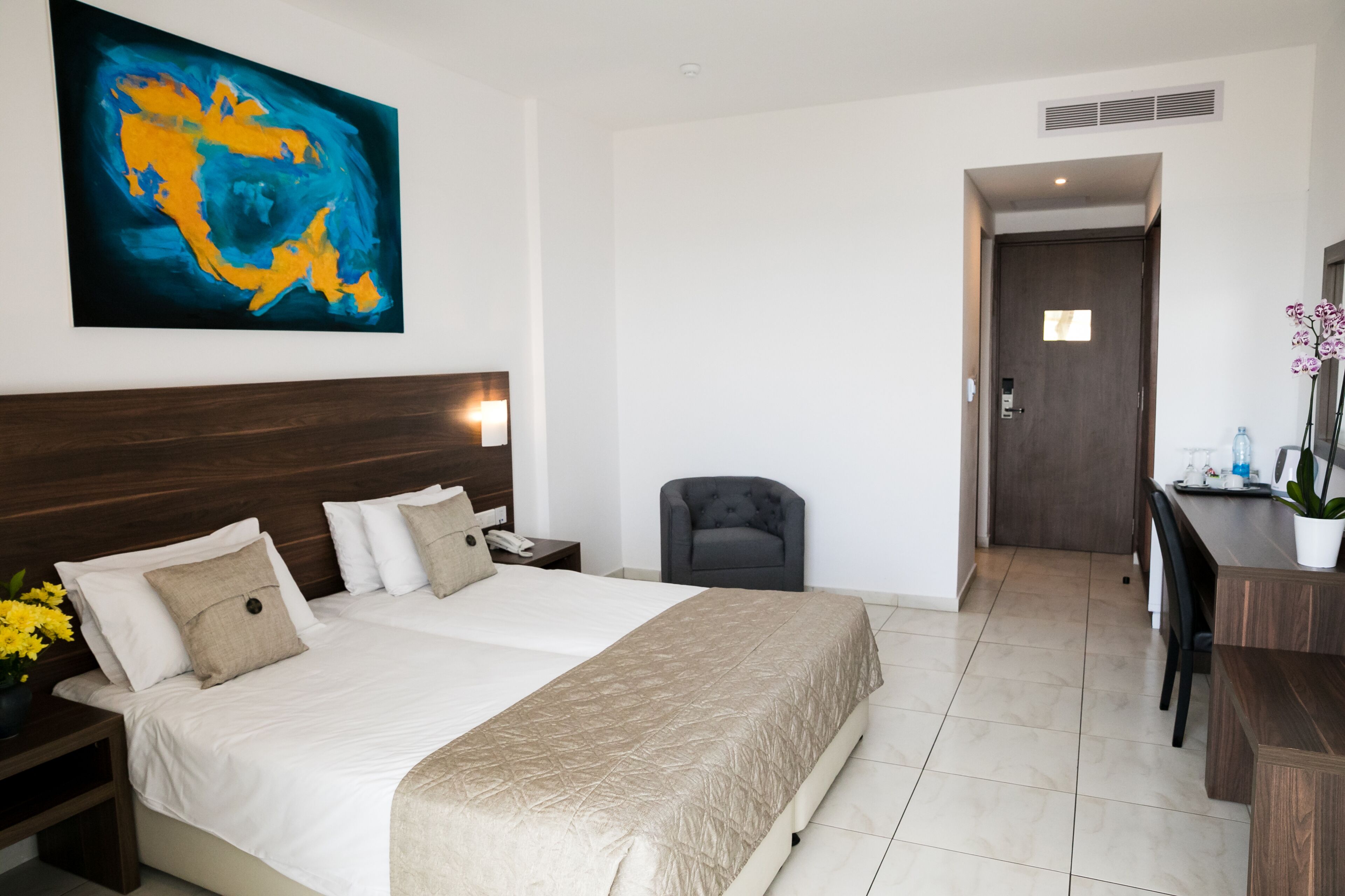 A hotel room with a large bed, a desk, a chair, a painting on the wall, and a door leading to another room.