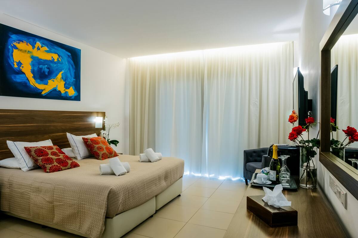 Une chambre d’hôtel avec un grand lit, une table de chevet avec une bouteille et des verres, une chaise, un grand miroir et un tableau au mur.