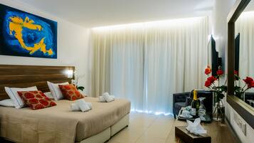 A hotel room with a large bed, a bedside table with a bottle and glasses, a chair, a large mirror, and a painting on the wall.