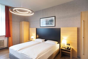 A modern hotel room with a large bed, bedside tables with lamps, a framed picture on the wall, and a wooden wardrobe.